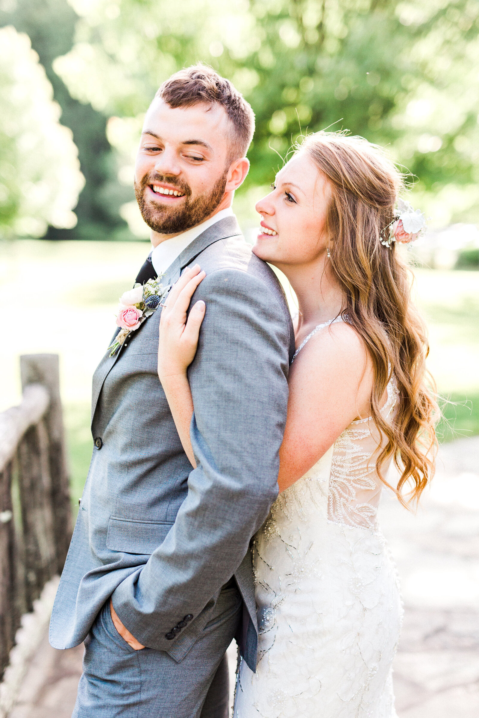 Bride embraces the groom from the side outdoors with a hand on his shoulder, both smiling, greenery behind them